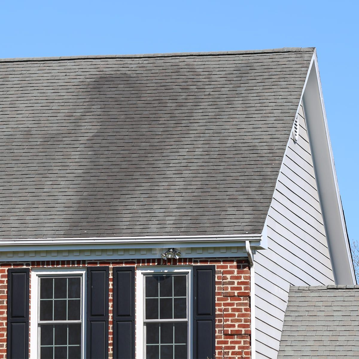 A shingled roof streaked with dark gloeocapsa magma algae above red brick and white trim.